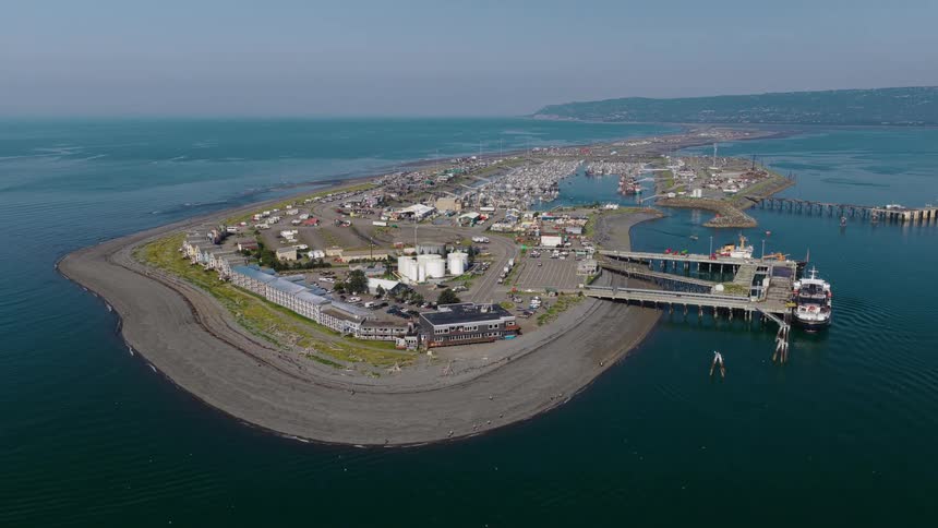A wide, static aerial view of the Homer Spit in Alaska, showcasing the bustling harbor, a docked ferry, and calm ocean waters on a sunny day