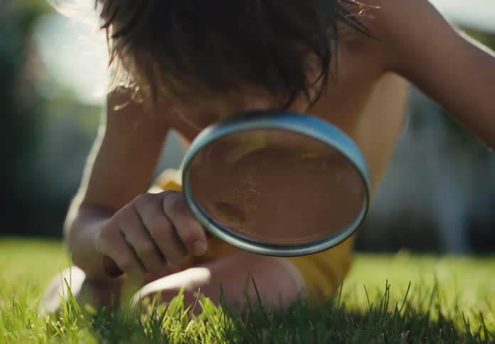 A curious young boy lies on a sunlit lawn, intently exploring the blades of grass with a magnifying glass
