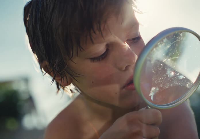 A curious young boy intently observes something through a magnifying glass with the bright sun flaring behind him