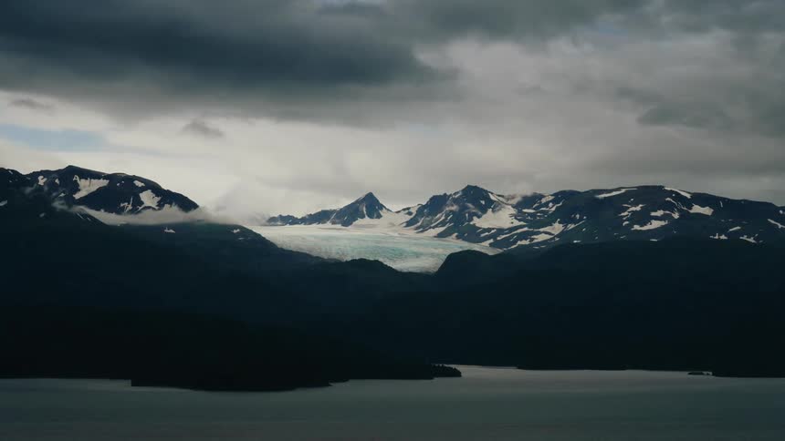 A dramatic time-lapse reveals dark, moody clouds rolling over a majestic glacier and snow-capped mountains