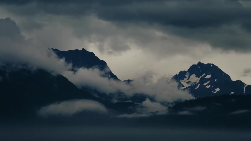 A dramatic time-lapse of dark storm clouds and fog rolling over snow-capped mountain peaks