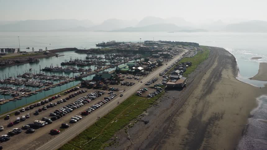 A high-angle aerial view slowly flies over the bustling Homer Spit in Alaska, with its harbor full of boats on one side and a sandy beach on the other