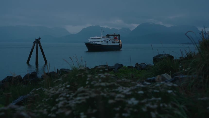 A ferry boat cruises through a tranquil bay with distant mountains under a dark, cloudy twilight sky
