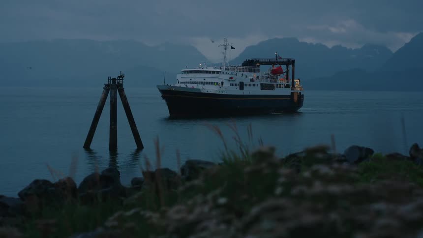 A ferry boat comes into focus as it sails on calm waters at dusk with mountains in the background