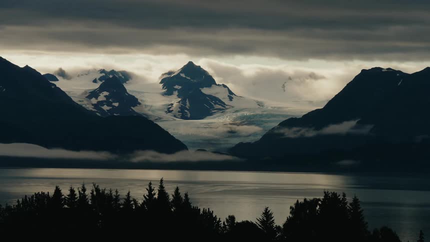 A stunning wide view of a majestic glacier between silhouetted mountains as clouds and fog roll over a calm lake