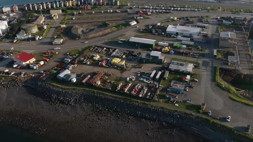 An aerial view slowly orbits an industrial harbor area with equipment yards and buildings along the coastline