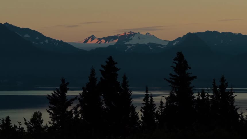 The setting sun illuminates the snowy peaks of distant mountains, viewed over a silhouetted forest and calm water in this serene landscape shot