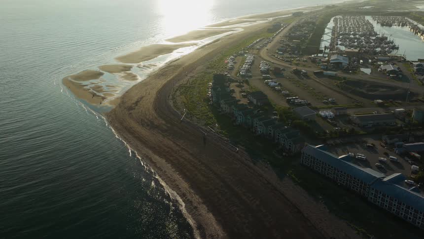 High aerial view captures a coastal town and marina nestled on a narrow spit of land as the sun sets over the calm ocean