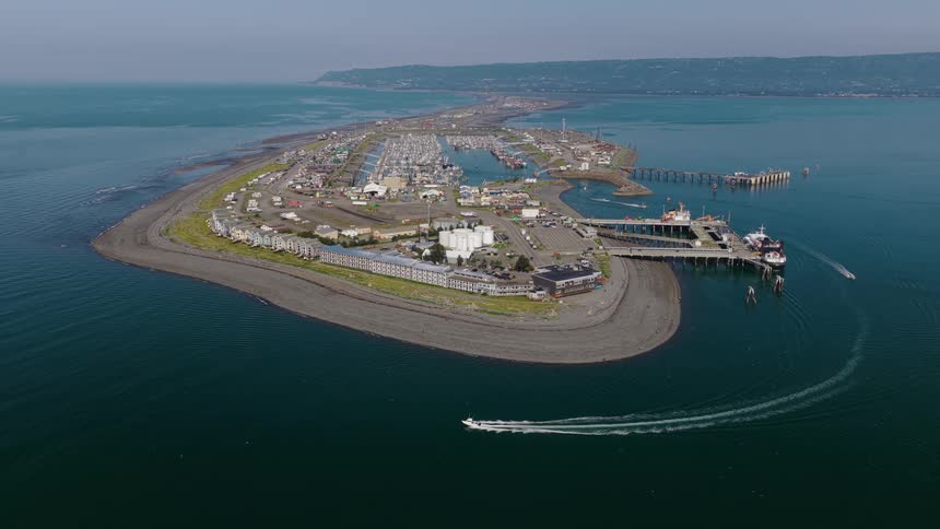 High aerial view of the Homer Spit in Alaska, a long peninsula with a marina, hotels, and an RV park, as a speedboat cruises by on a calm day