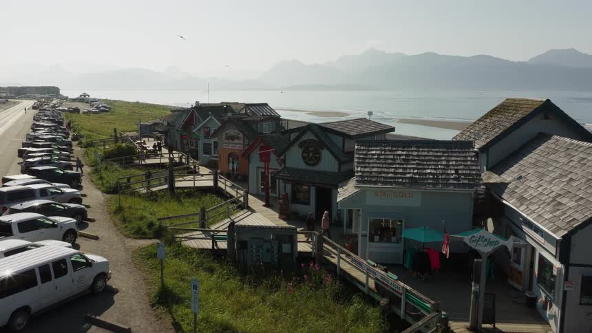 An aerial view slowly moves over a quaint coastal boardwalk village with shops and tourists next to a beach and mountains in the background