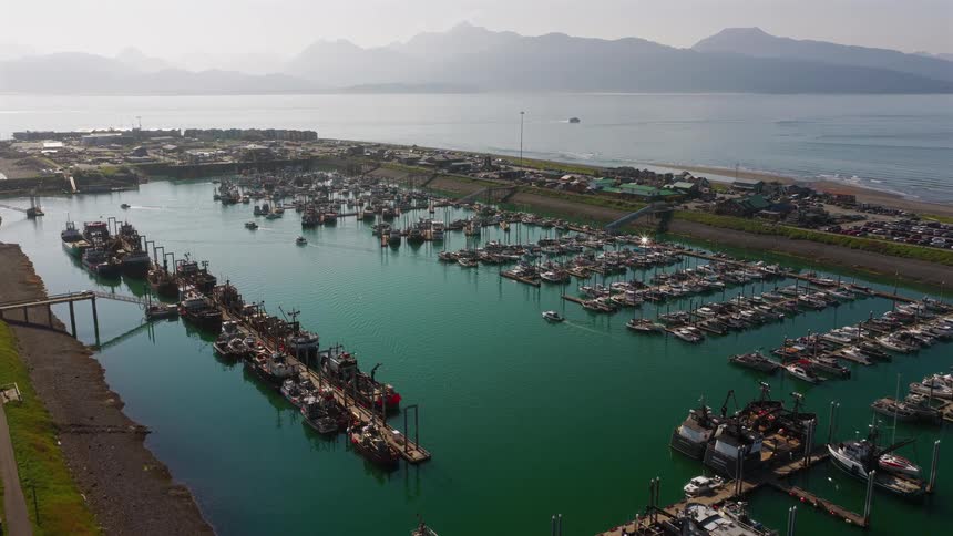A slow aerial push-in shot reveals a large, busy harbor filled with fishing boats, with the ocean and misty mountains in the background