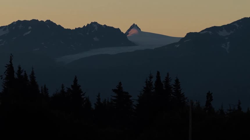A majestic mountain peak catches the last light of the sunset, glowing brightly above a silhouetted forest and glacier