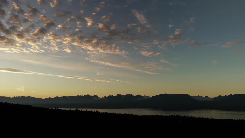 A serene sunrise illuminates scattered clouds above a silhouetted mountain range and a vast, calm lake