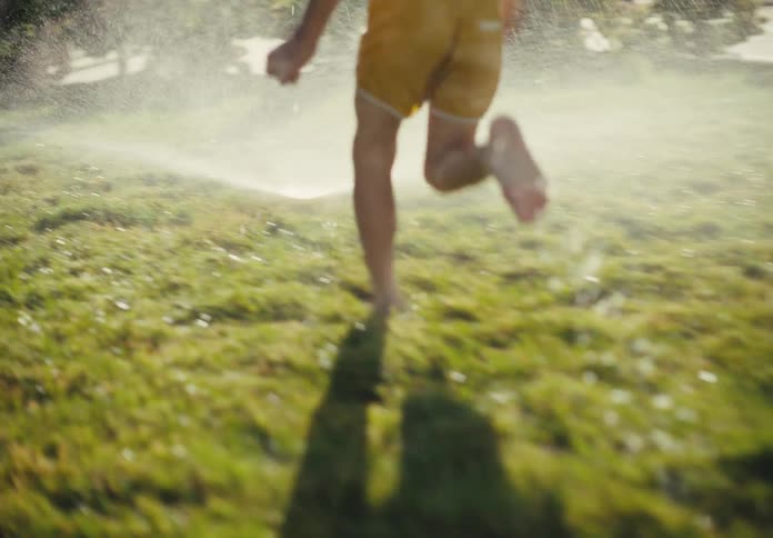 A barefoot boy in yellow shorts runs through a lawn sprinkler on a lush green lawn in slow motion