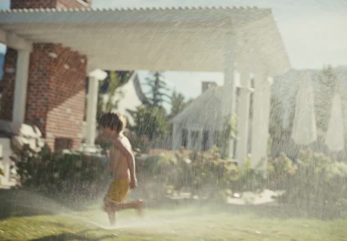 A happy shirtless boy in yellow shorts runs through a sprinkler in slow motion in a sun-drenched backyard