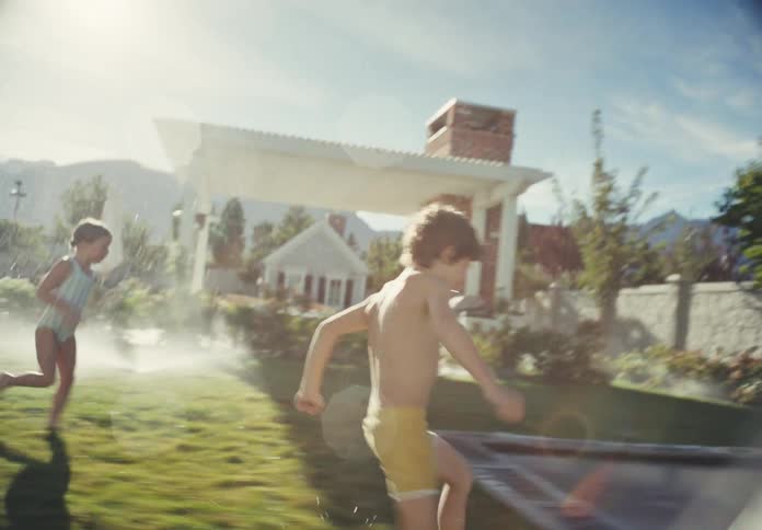 A young boy and girl in swimsuits joyfully run through a sprinkler and jump on a trampoline in their backyard