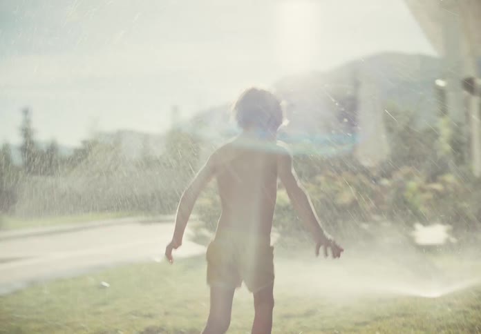 A young shirtless boy runs through a sprinkler in slow motion on the lawn of a suburban house on a sunny day
