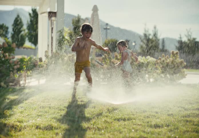 Two carefree young siblings laugh and play while running through a sprinkler in their sun-drenched backyard in slow motion