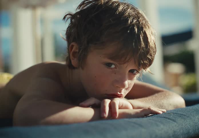 A thoughtful young boy with wet hair rests on a lounge chair, looking directly into the camera with a pensive expression
