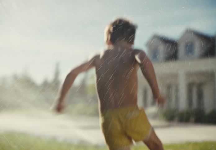 A joyful young boy in yellow shorts runs through a lawn sprinkler in slow motion on a bright, sunny summer day in his backyard