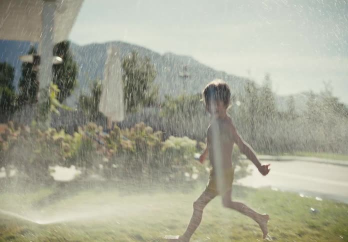 Carefree children laugh and run through a lawn sprinkler on a sunny summer day in slow motion