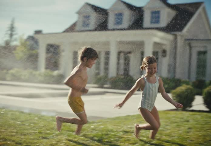 A young boy and girl in swimsuits laugh and run through a sprinkler on a grassy lawn in slow motion on a sunny summer day