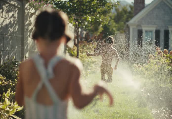 Over-the-shoulder slow motion shot of a young boy and girl running through a backyard sprinkler on a sunny summer day