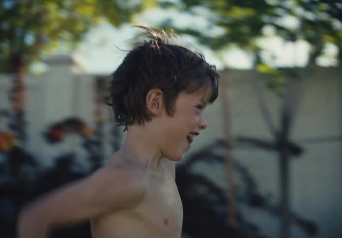 Shirtless young boy runs playfully through a sunny backyard garden with motion blur and summer energy