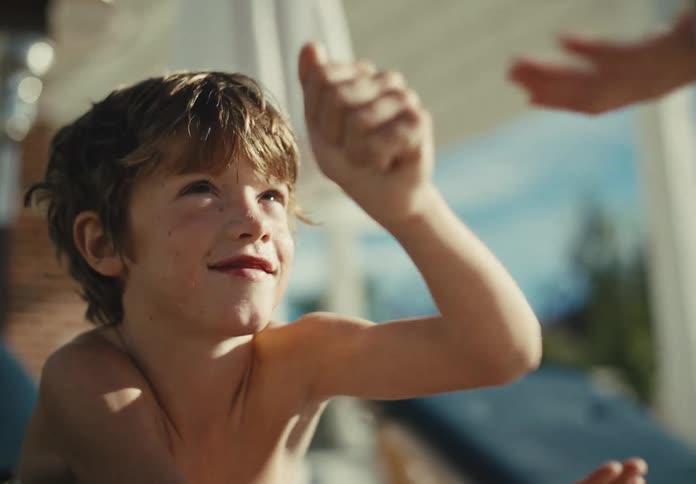Smiling young boy relaxes shirtless on a sunny lounge chair and reaches up for a playful hand interaction outdoors