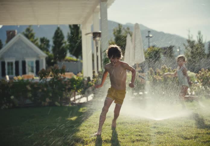 Carefree kids run and play in a backyard sprinkler during a sunny summer afternoon in slow motion