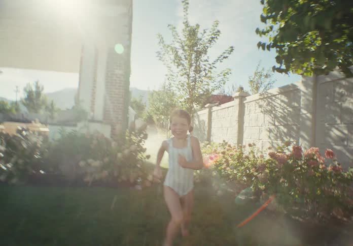Smiling little girl in a swimsuit joyfully runs through the backyard sprinklers on a sunny summer day in slow motion