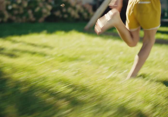A carefree boy in yellow shorts runs barefoot across a sunlit lawn, kicking a soccer ball and splashing through water in slow motion