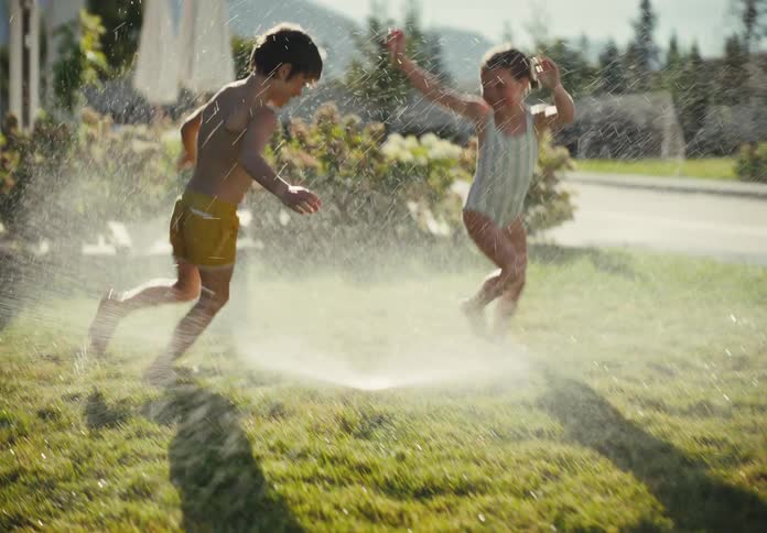 Two happy children, a boy and a girl, laugh and run through a backyard sprinkler in slow motion on a sunny summer day