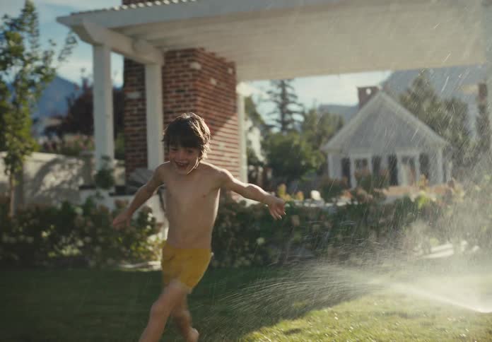 A happy young boy runs through a lawn sprinkler with his arms outstretched on a sunny summer day in slow motion