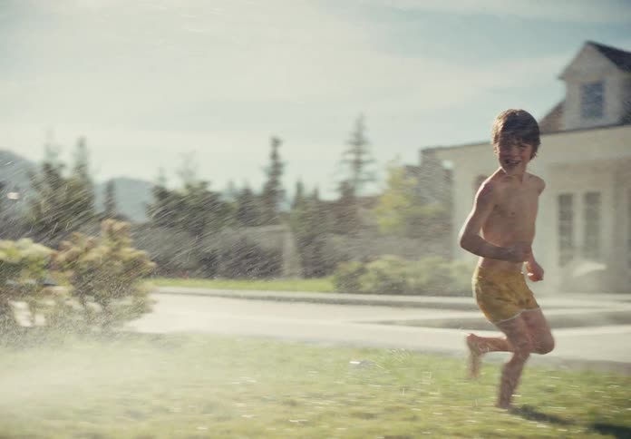 Happy kids run barefoot through a backyard sprinkler in slow motion on a sunny summer day