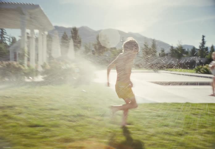 Slow motion shot of a young boy playfully running through the sprinklers in a sun-drenched backyard