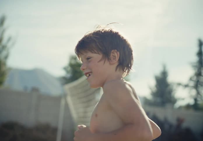 Shirtless young boy runs barefoot across a sunny backyard lawn near a soccer goal
