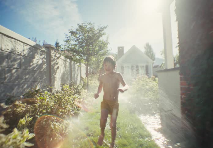 A joyful young boy in swim trunks runs towards the camera in slow motion through a backyard sprinkler on a bright summer day