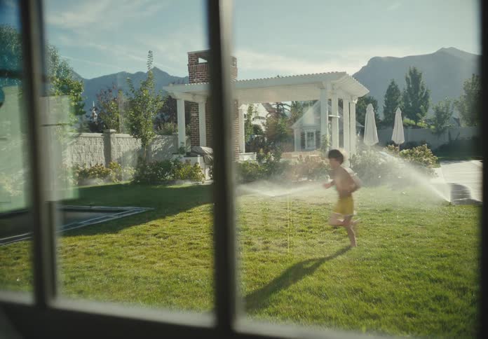 Slow motion view through a window of two kids gleefully running through backyard sprinklers on a sunny day