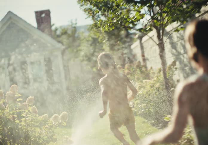 Slow motion shot of two happy children running and playing in a sprinkler in a sun-drenched backyard