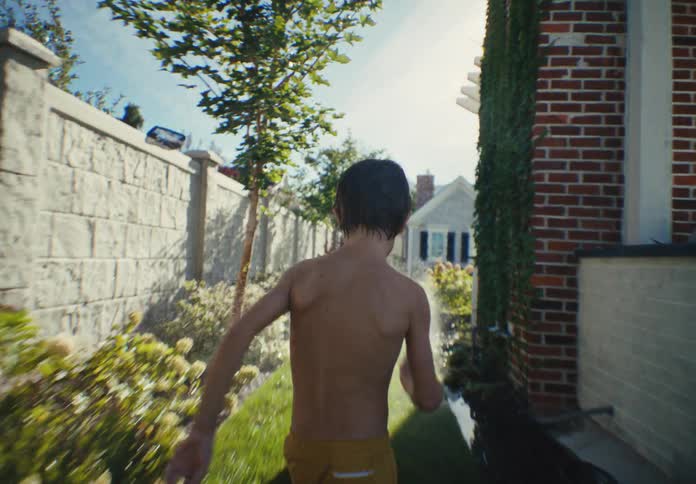 A slow-motion following shot captures two children running barefoot through the sprinklers in a sun-drenched backyard
