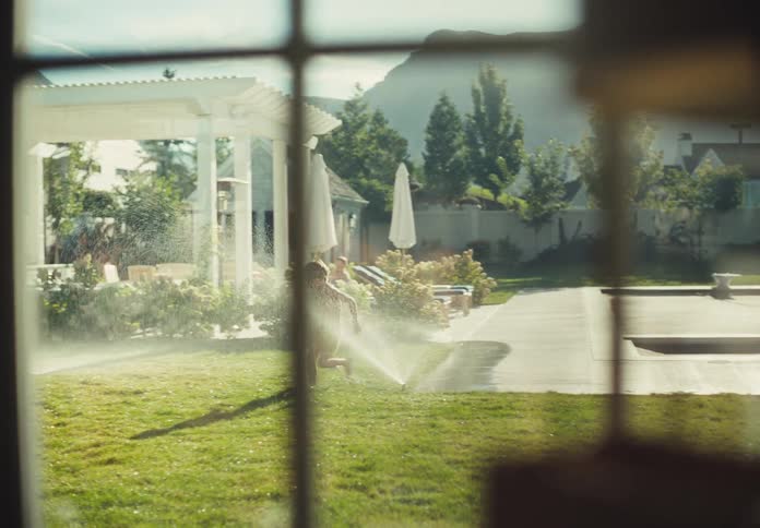 A slow-motion shot from a window captures a carefree boy running through sprinklers in a sunny suburban backyard