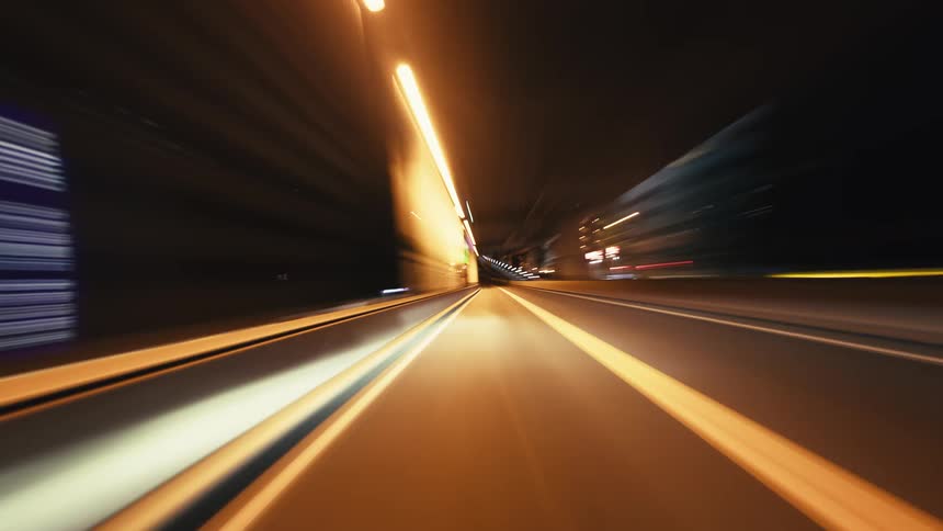 A dynamic first-person point-of-view time-lapse shows a car speeding through an illuminated urban highway tunnel at night, creating mesmerizing light streaks