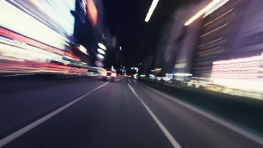 A dizzying point-of-view hyperlapse shows a car speeding through a neon-lit city at night, creating vibrant light trails