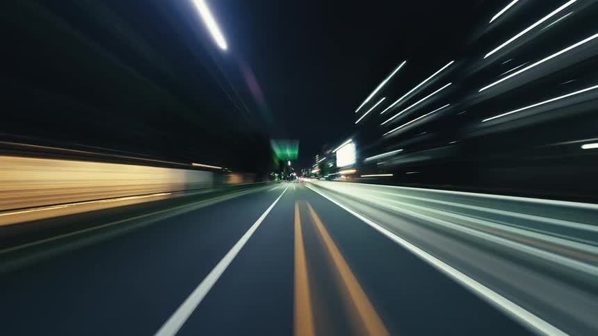A first-person point-of-view hyper-lapse captures a high-speed drive through a city at night, with buildings and traffic creating vibrant light streaks
