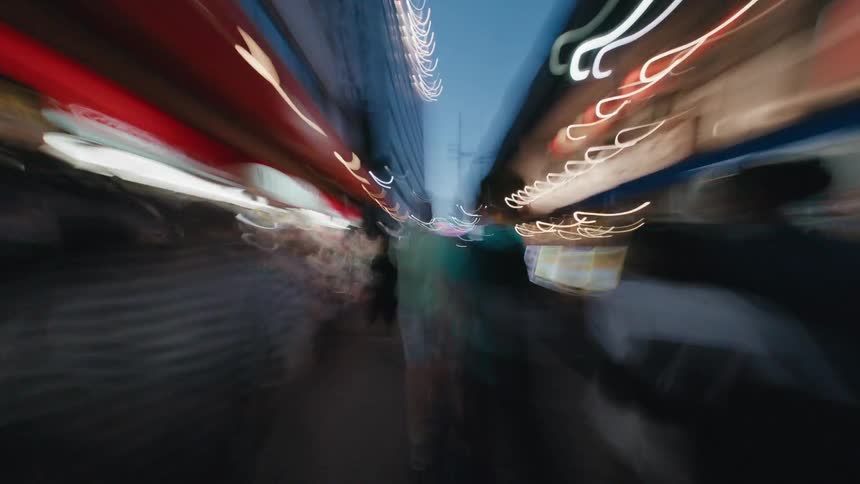 A fast-paced POV hyperlapse rushes through a bustling and crowded city market street at dusk, with lights blurring into vibrant streaks