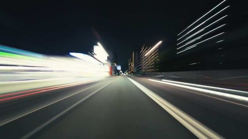 A dynamic point-of-view hyperlapse shows a car speeding through city streets at night, transforming lights into vibrant streaks of color