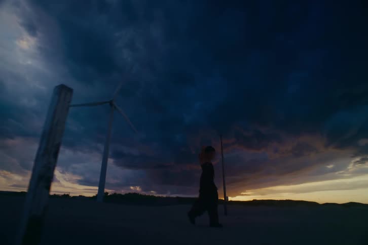 In slow motion, a woman walks and dances in a desolate landscape with a wind turbine under a dramatic stormy sky at sunset