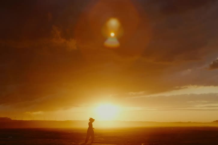 A silhouette of a woman walks through a vast, open landscape under a dramatic golden sky at sunset