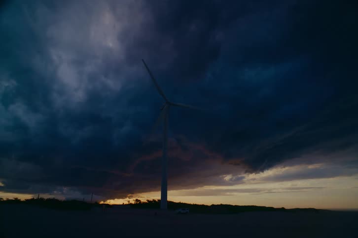 A lone wind turbine turns against a dark, dramatic sky as lightning flashes within the storm clouds at dusk
